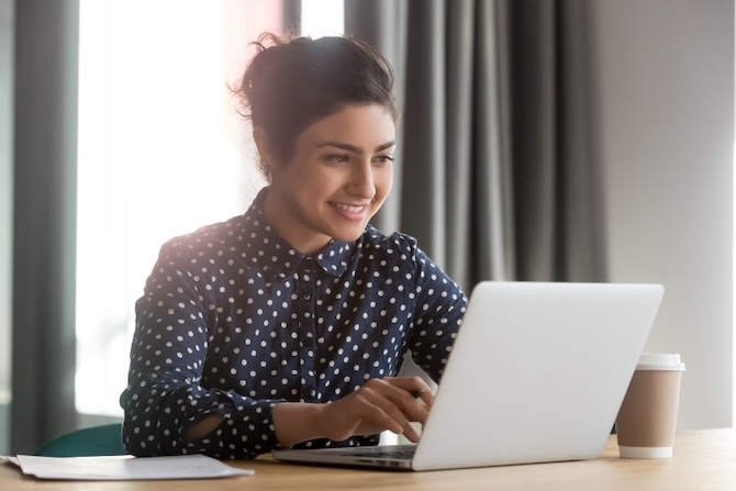 Picture of a woman using a laptop sat at a desk near a window.
